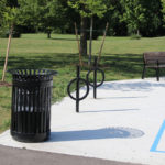 Une poubelle noire, deux porte-vélos circulaires et un banc sont posés sur une aire bétonnée dans un parc ensoleillé avec de l'herbe verte et des arbres.
