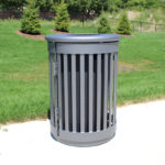 A cylindrical metal trash can with vertical slats stands on a concrete path in a grassy park area with trees and a fence in the background.
