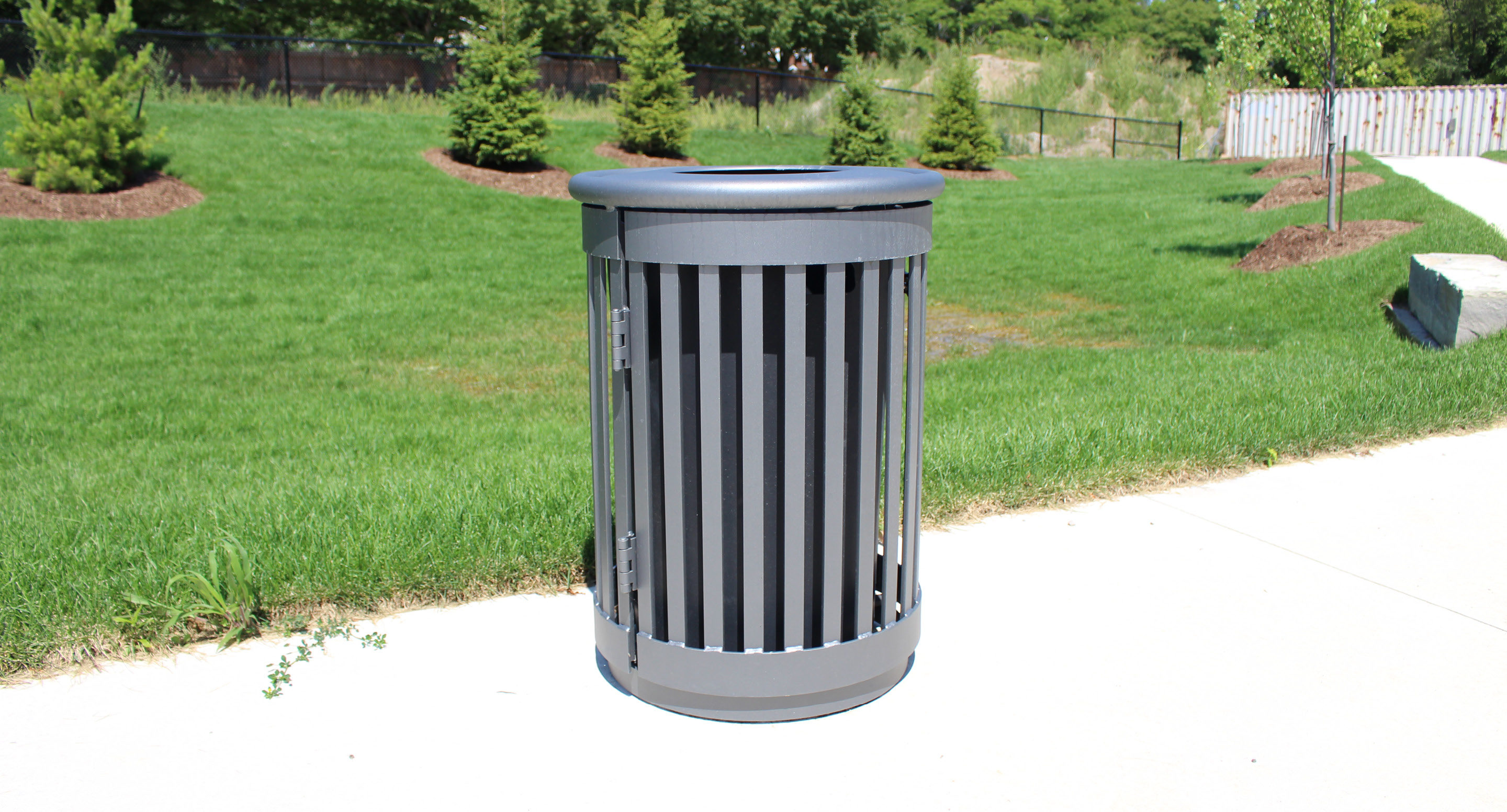 maglin_mtr-0250-trash-receptacles-0611 A cylindrical metal trash can with vertical slats stands on a concrete path in a grassy park area with trees and a fence in the background.