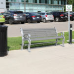 Two metal benches and a trash can on a sidewalk near a parking lot with parked cars and a building in the background.