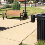 A black metal trash can stands near two wooden benches on a concrete walkway in a landscaped park area with grass, shrubs, and parked cars in the background.