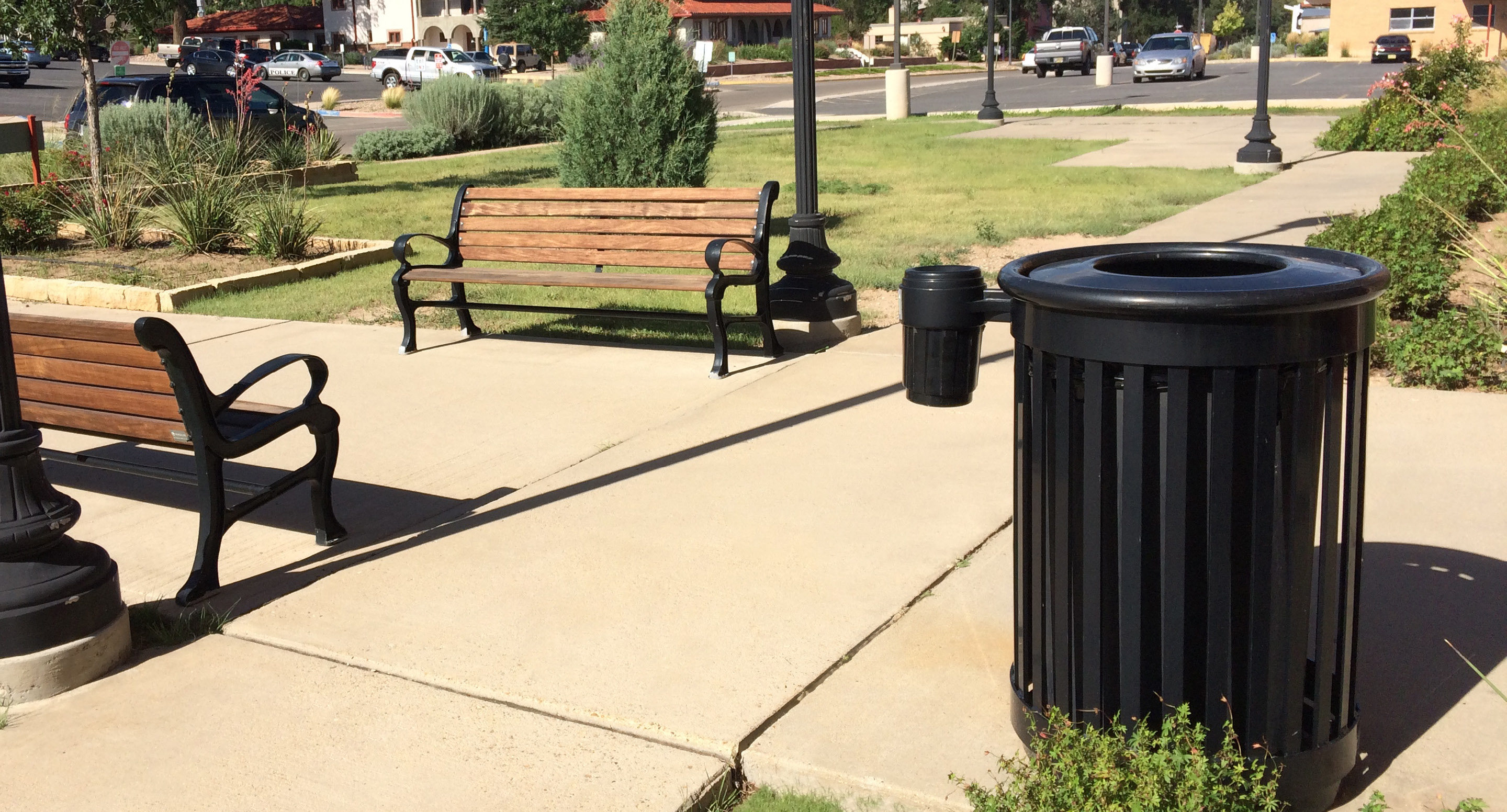 maglin_mtr-0250-trash-receptacles-2 A black metal trash can stands near two wooden benches on a concrete walkway in a landscaped park area with grass, shrubs, and parked cars in the background.