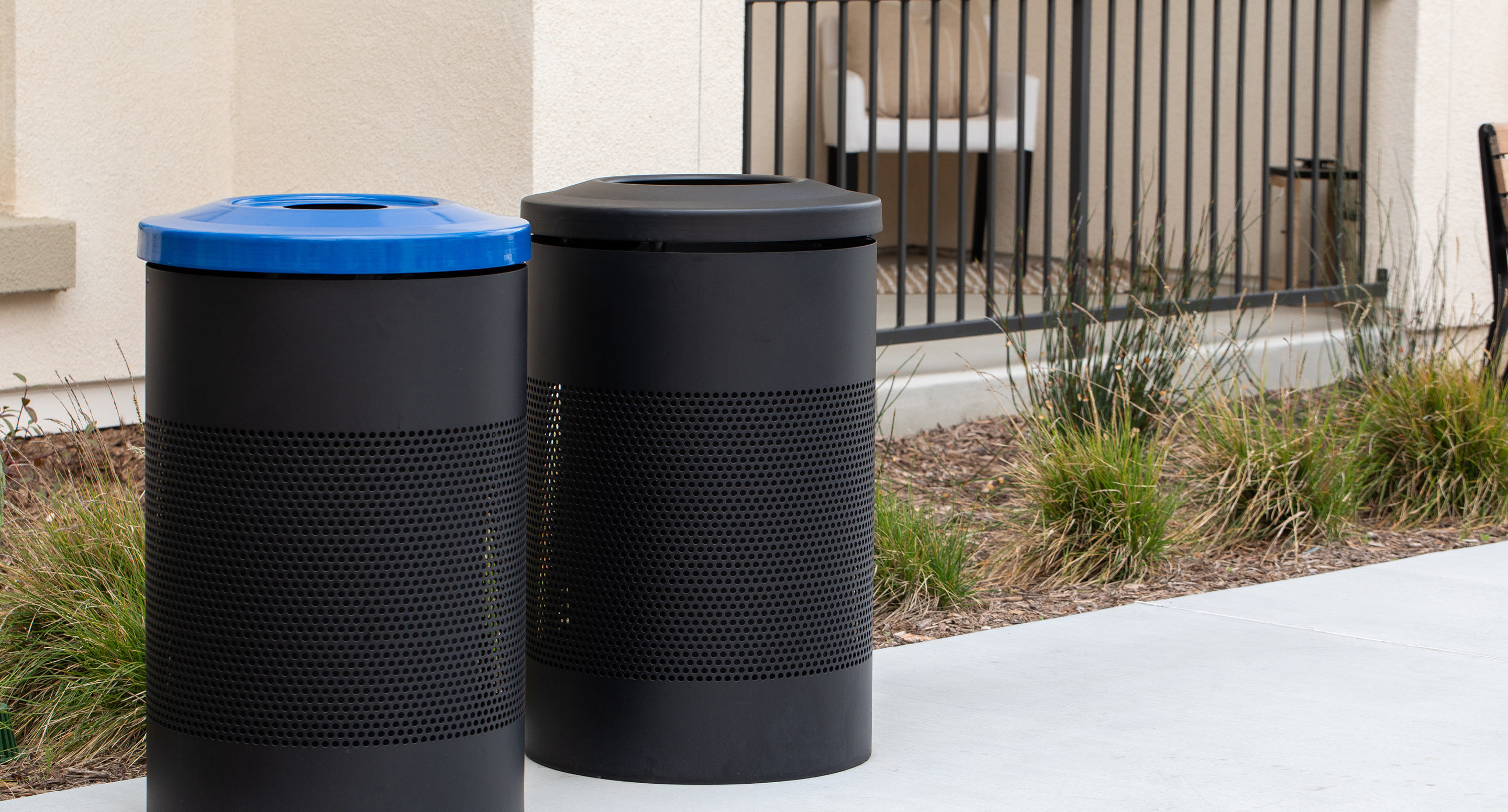 maglin_mtr-0600-trash-receptacles-2 Two black outdoor trash bins stand side by side on a sidewalk; one has a blue lid, and landscaping with grass is visible behind them.