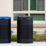 Two black cylindrical outdoor trash bins, one with a blue lid and one with a black lid, are placed on a sidewalk in front of a building with a window and greenery.