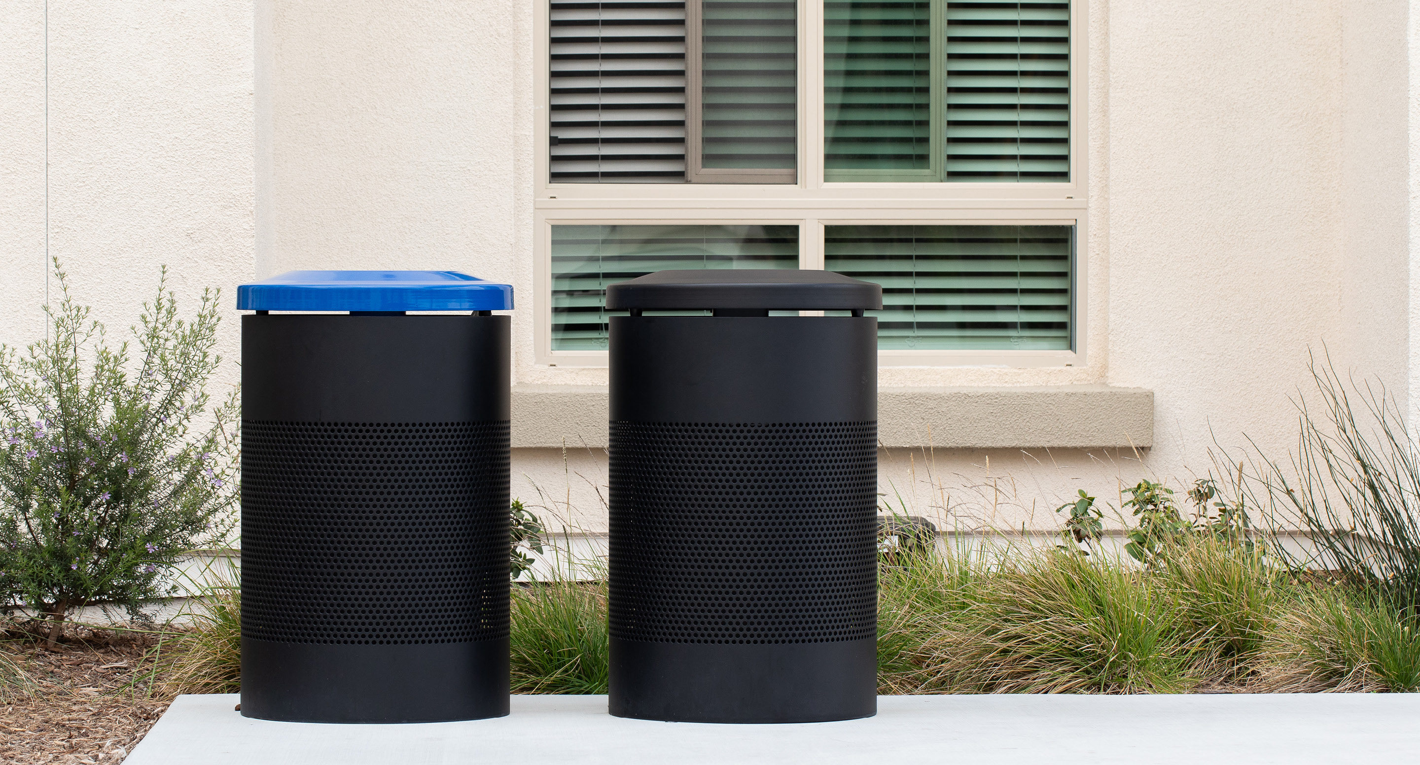 maglin_mtr-0600-trash-receptacles-3 Two black cylindrical outdoor trash bins, one with a blue lid and one with a black lid, are placed on a sidewalk in front of a building with a window and greenery.