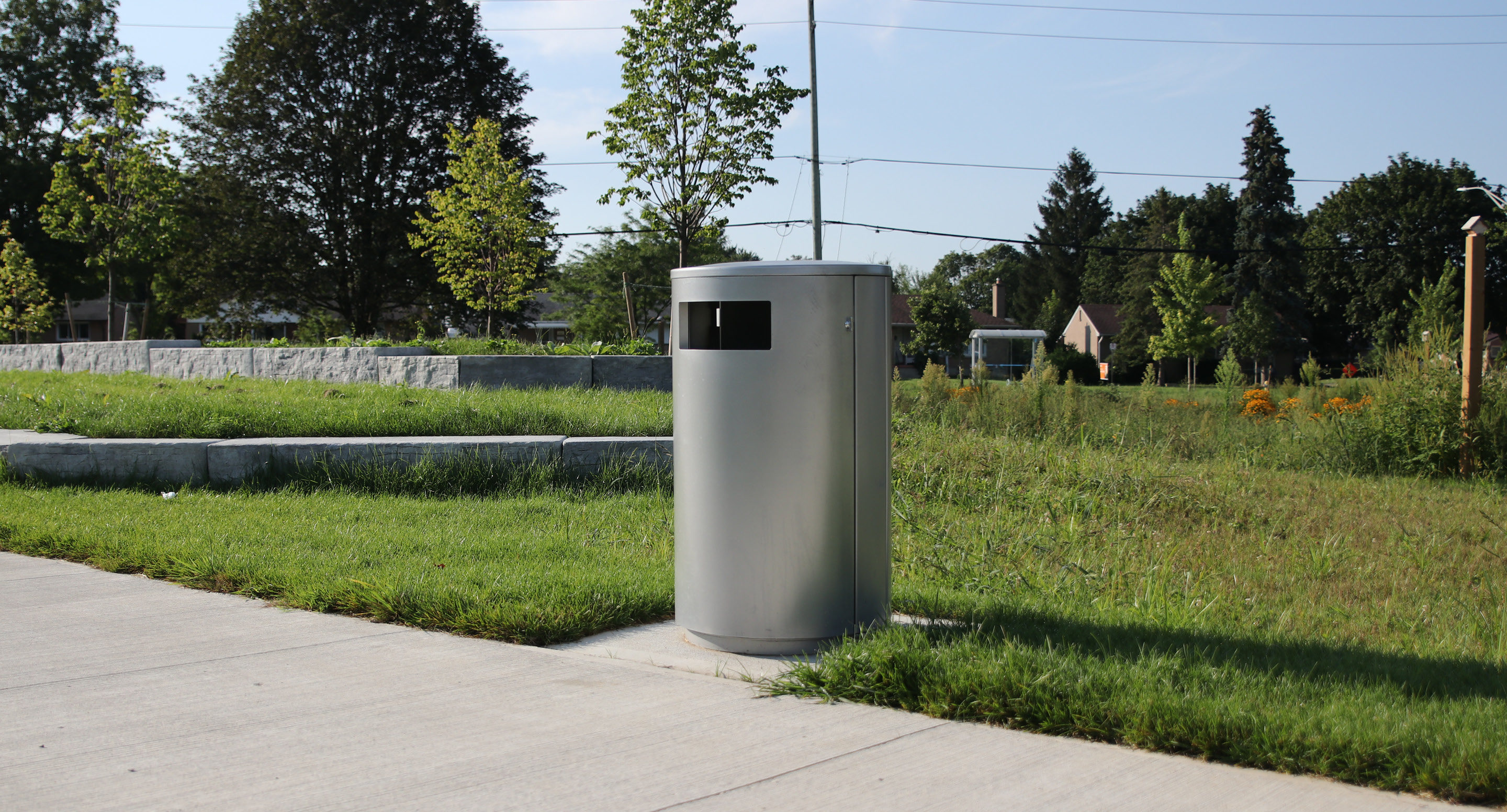 maglin_mtr-0650-trash-receptacles-3-0613 Une poubelle en métal argenté est placée sur un trottoir en béton à côté d'une zone herbeuse avec des arbres et des murets en pierre à l'arrière-plan.