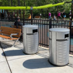 Deux poubelles argentées étiquetées "Recycle" et "Trash" se trouvent près d'un banc à l'extérieur d'une aire de jeux clôturée où se trouvent des enfants et des adultes.