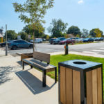 Un banc public et une poubelle sont situés sur un trottoir près d'un parking avec plusieurs voitures. Des arbres, un passage piéton et un clocher d'église sont visibles à l'arrière-plan.