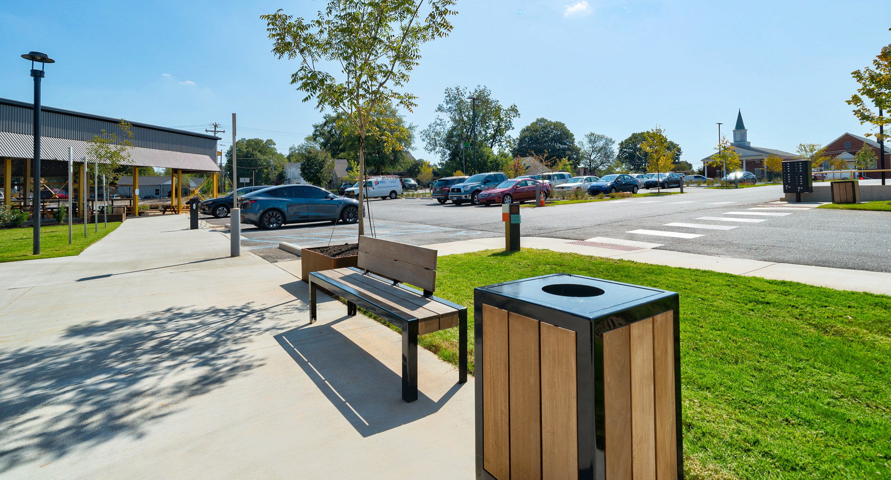 Un banc public et une poubelle sont situés sur un trottoir près d'un parking avec plusieurs voitures. Des arbres, un passage piéton et un clocher d'église sont visibles à l'arrière-plan.