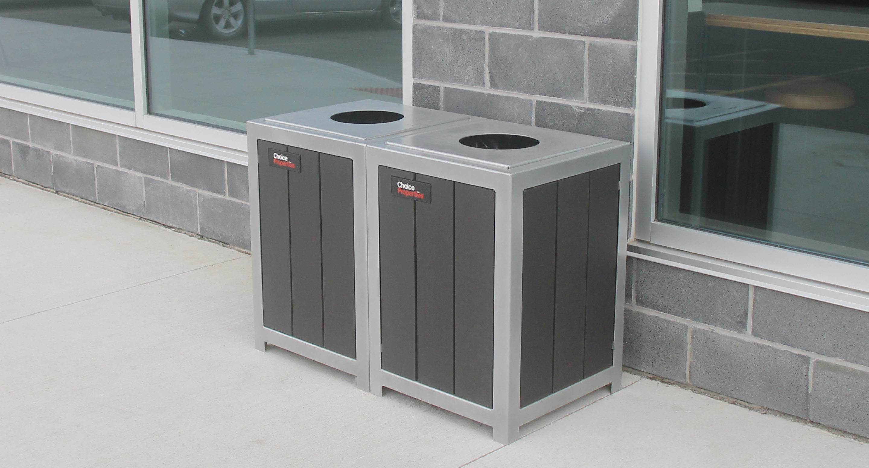 maglin_mtr-1050-trash-receptacles-2 Deux poubelles noires et argentées sont placées côte à côte contre un mur de briques grises à l'extérieur d'un bâtiment aux grandes fenêtres.