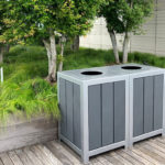 Deux poubelles grises d'extérieur se trouvent sur une terrasse en bois à côté d'une jardinière surélevée avec de l'herbe verte et de petits arbres.