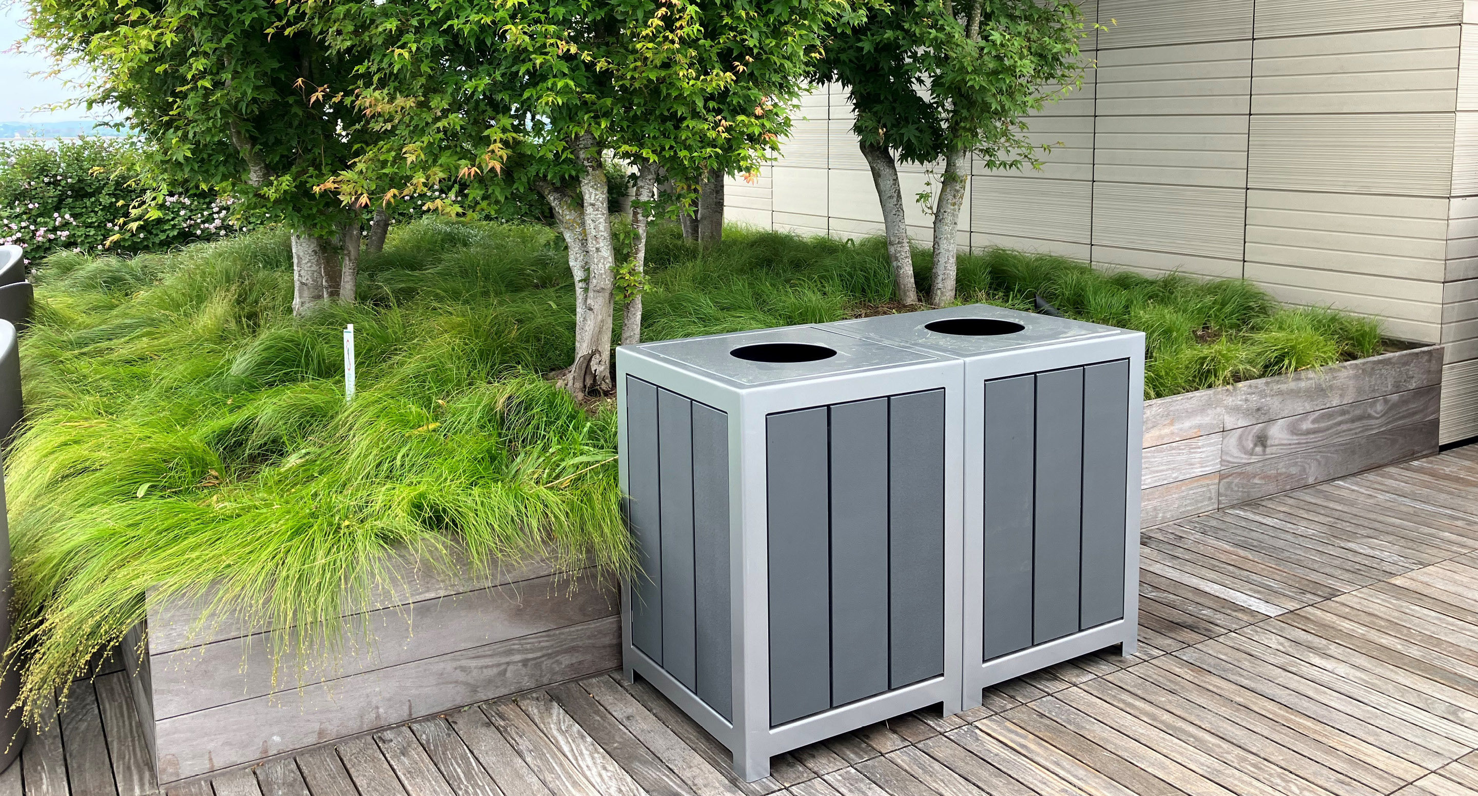 maglin_mtr-1050-trash-receptacles-3 Deux poubelles grises d'extérieur se trouvent sur une terrasse en bois à côté d'une jardinière surélevée avec de l'herbe verte et de petits arbres.