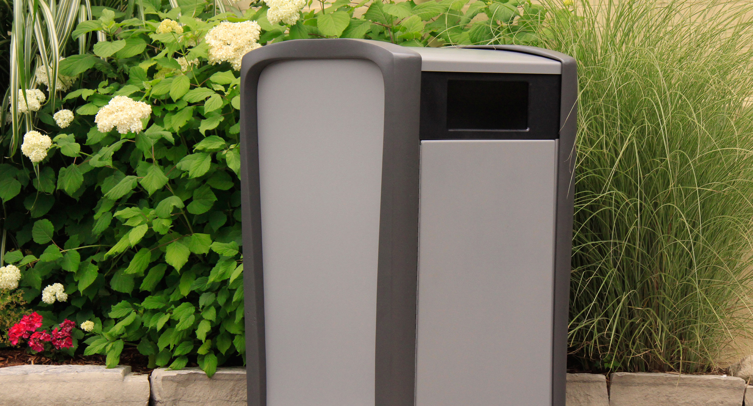 maglin_mtr-1400-trash-receptacles-1-0613b Une poubelle d'extérieur grise et noire est placée sur un trottoir en pierre devant des plantes vertes et des fleurs blanches.