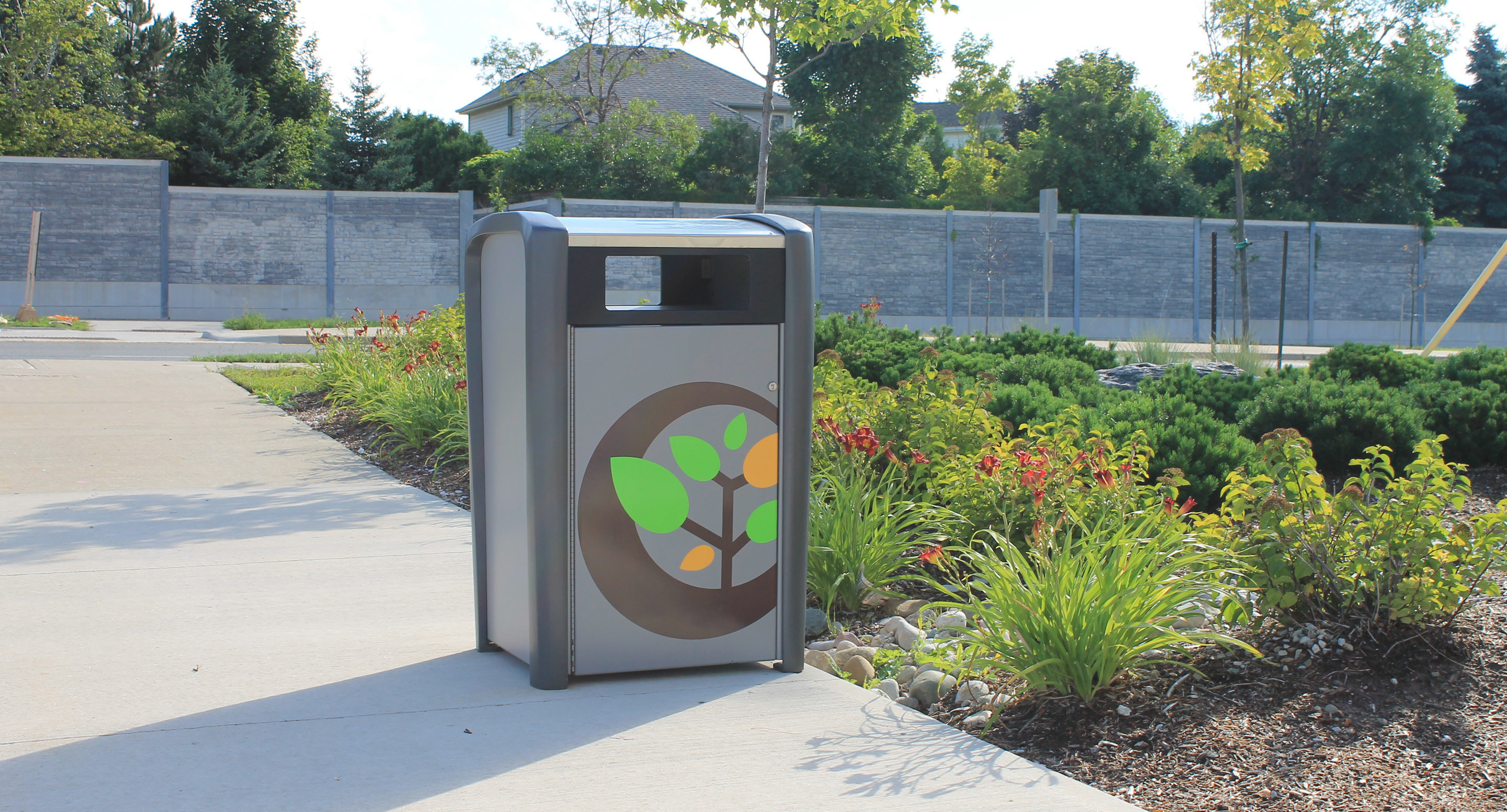 maglin_mtr-1400-trash-receptacles-3-0613b Une poubelle ornée d'un motif de feuilles se trouve sur un trottoir, à côté d'un espace vert aménagé et de fleurs dans un cadre extérieur.