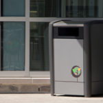 Une poubelle extérieure grise portant un logo de recyclage se trouve à côté d'un bâtiment doté de grandes baies vitrées et d'un mur d'accent rouge.