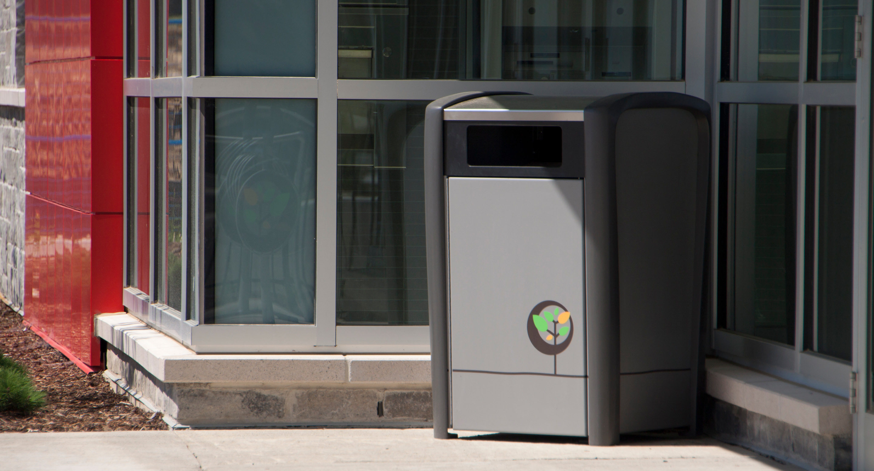 Une poubelle extérieure grise portant un logo de recyclage se trouve à côté d'un bâtiment doté de grandes baies vitrées et d'un mur d'accent rouge.