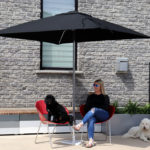 A woman sits on a red chair under a black patio umbrella with two white poodles, on a concrete patio outside a brick building.