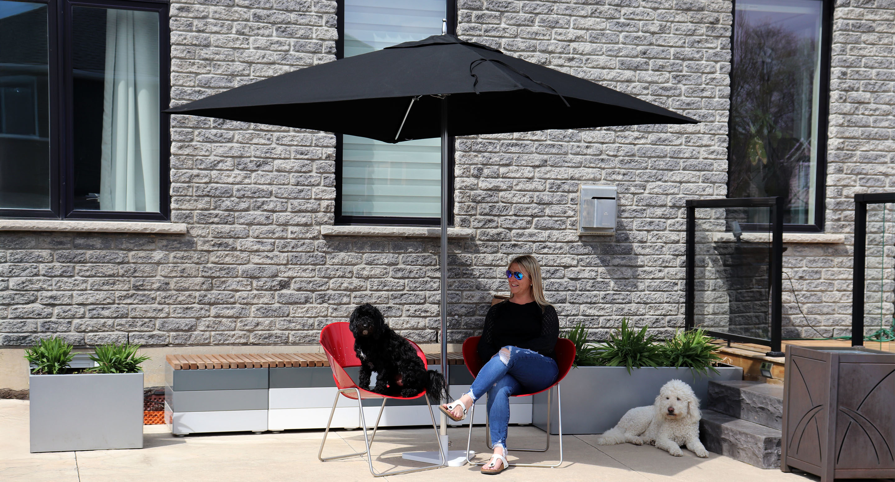 maglin_mum-2400-tuuci-umbrella-1 A woman sits on a red chair under a black patio umbrella with two white poodles, on a concrete patio outside a brick building.