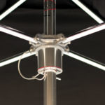 Close-up view of the underside of a large umbrella, showing the central pole, metal spokes, and attached wiring against a dark background.