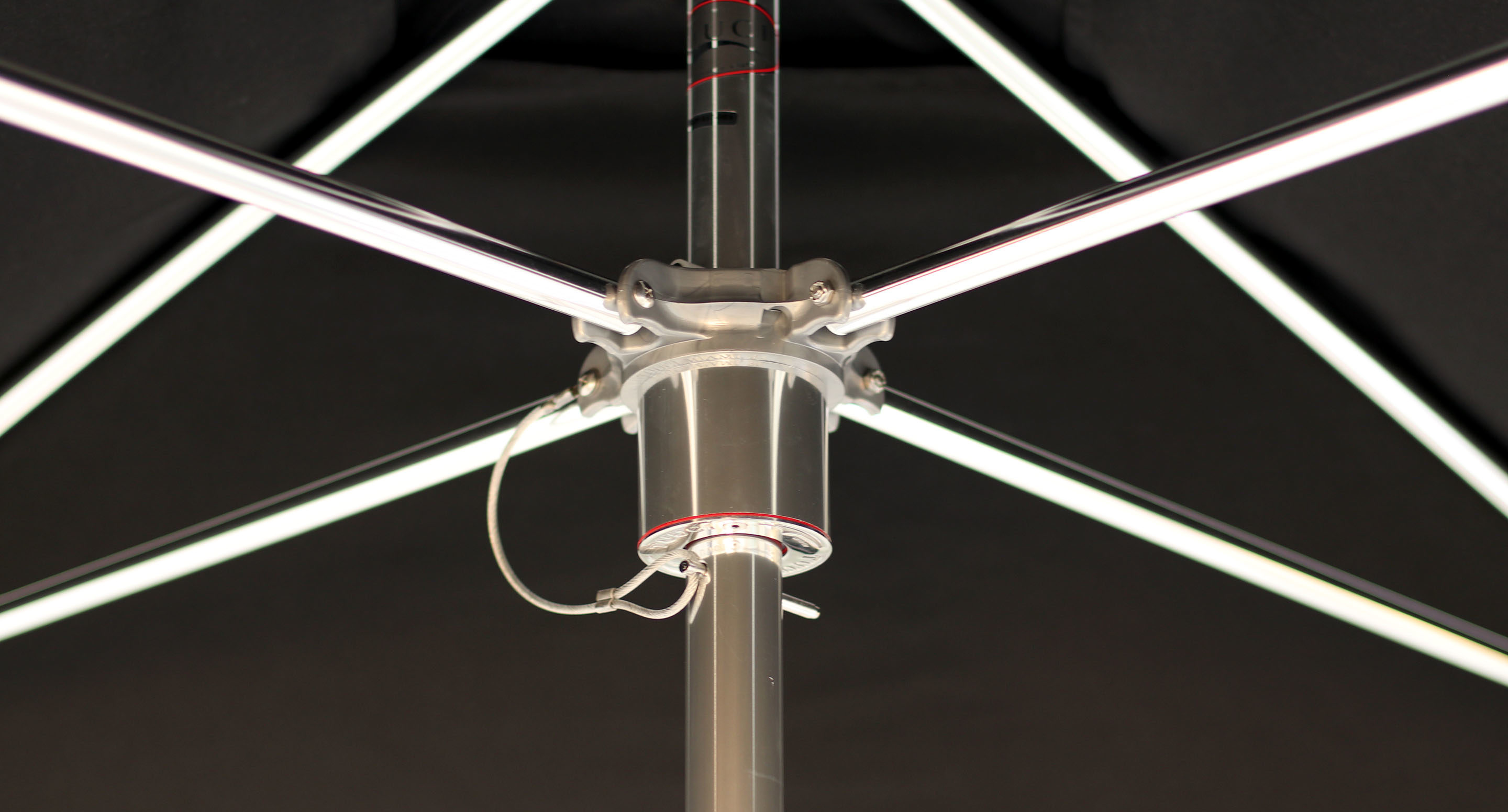 maglin_mum-2400-tuuci-umbrella-2 Close-up view of the underside of a large umbrella, showing the central pole, metal spokes, and attached wiring against a dark background.