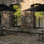 Two round tables with metal chairs and black umbrellas are set on a cobblestone patio next to a stone wall with arched openings and iron railings, facing a scenic outdoor area.