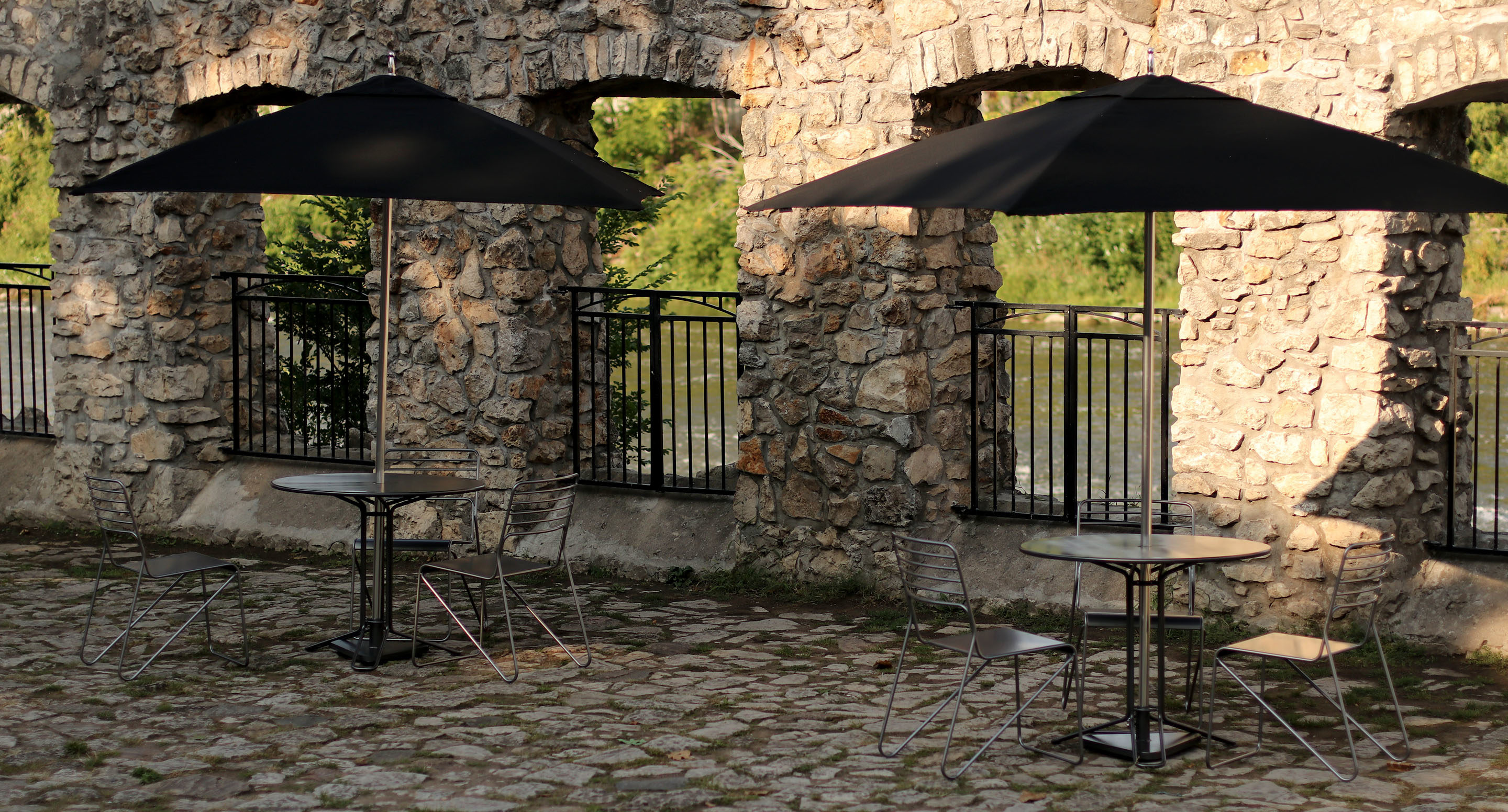 maglin_mum-2400-tuuci-umbrella-3 Two round tables with metal chairs and black umbrellas are set on a cobblestone patio next to a stone wall with arched openings and iron railings, facing a scenic outdoor area.