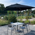 A square table with four chairs sits under a large black umbrella on a gravel surface, with a park and playground in the background.