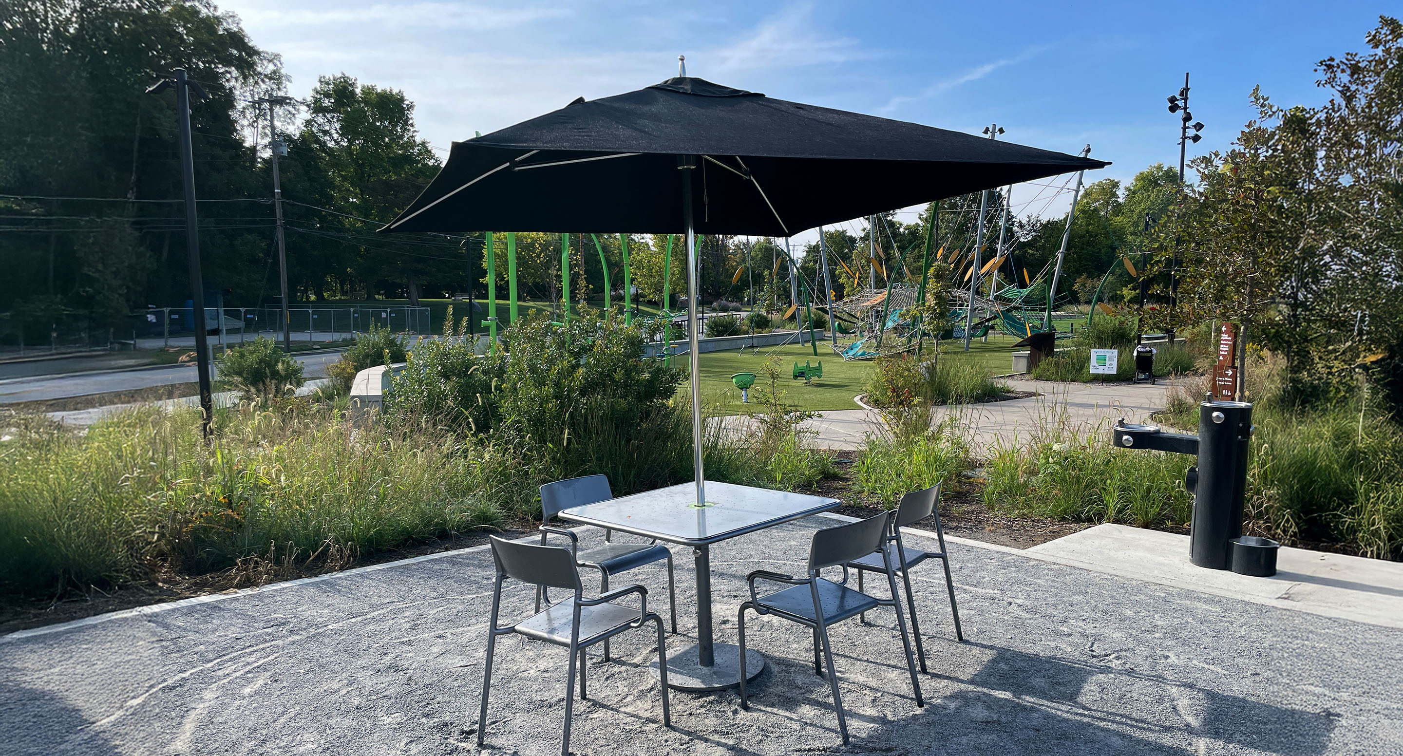 maglin_mum-2400-tuuci-umbrella-4 A square table with four chairs sits under a large black umbrella on a gravel surface, with a park and playground in the background.