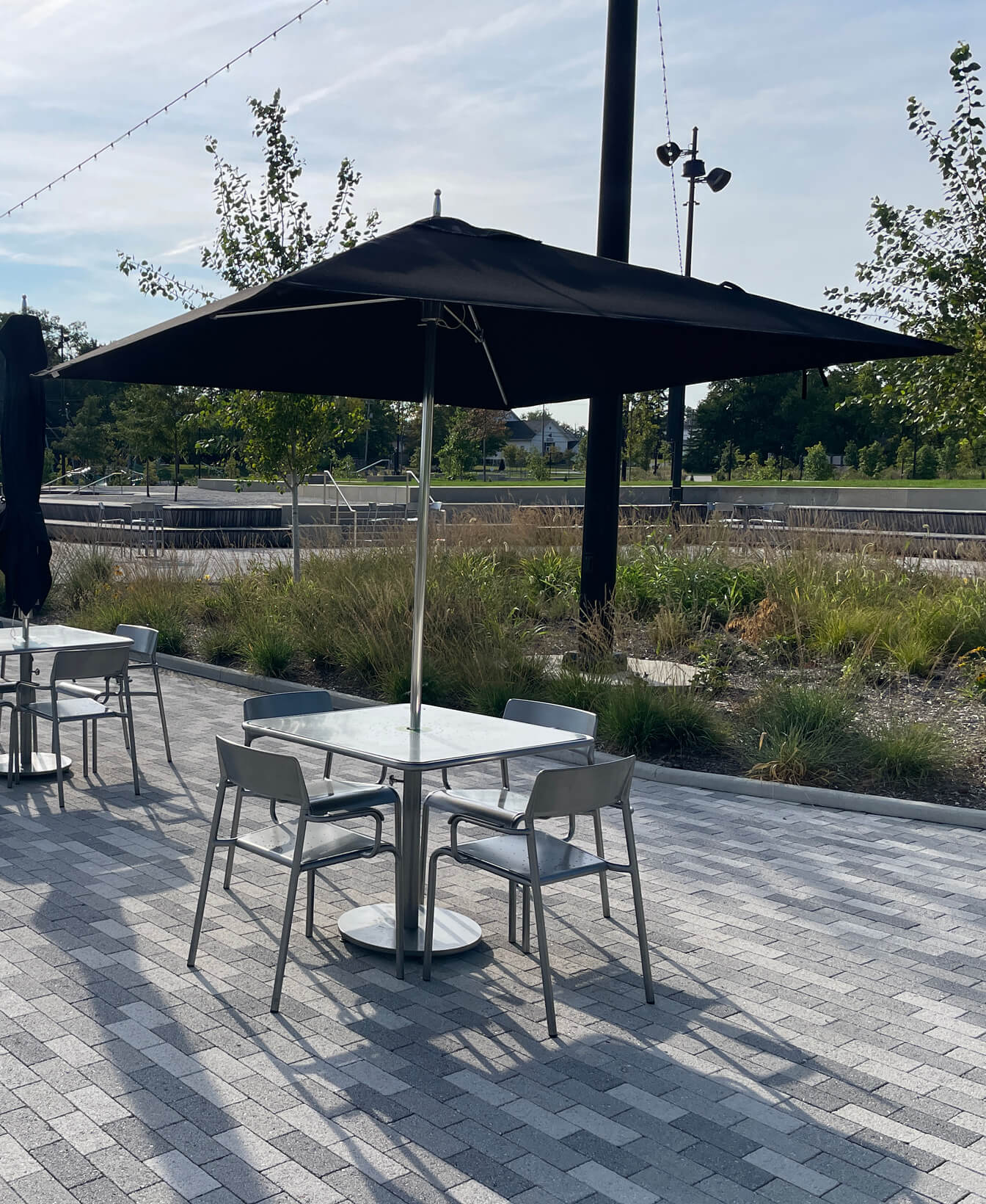 A metal outdoor table with four chairs and a large TUUCI Ocean Master Square Umbrella is set on a paved patio near landscaped greenery and trees.