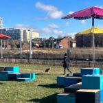 Des bancs géométriques colorés avec de grands parasols sont posés sur l'herbe près d'un chemin ; une statue et un chien sont visibles, avec des bâtiments de la ville à l'arrière-plan.