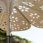 Gros plan d'un parapluie blanc en métal avec des motifs géométriques découpés, vu de dessous sur un fond flou en extérieur.