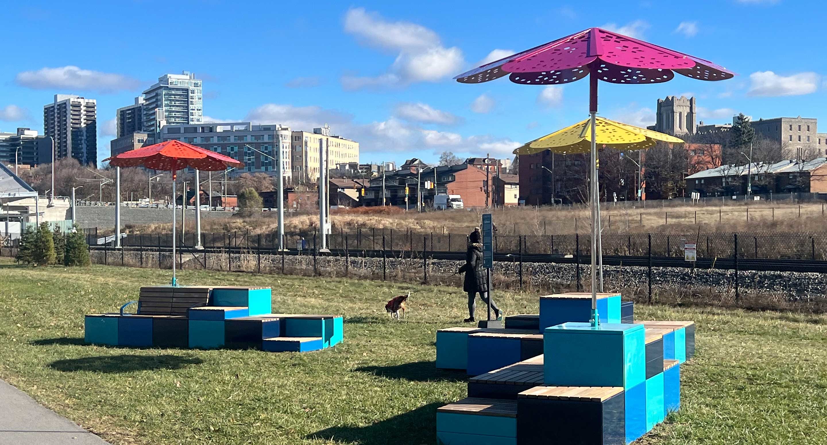 Colorful geometric benches with large metal umbrellas stand on grass near a path, with buildings, a statue, a dog, and blue sky in the background.