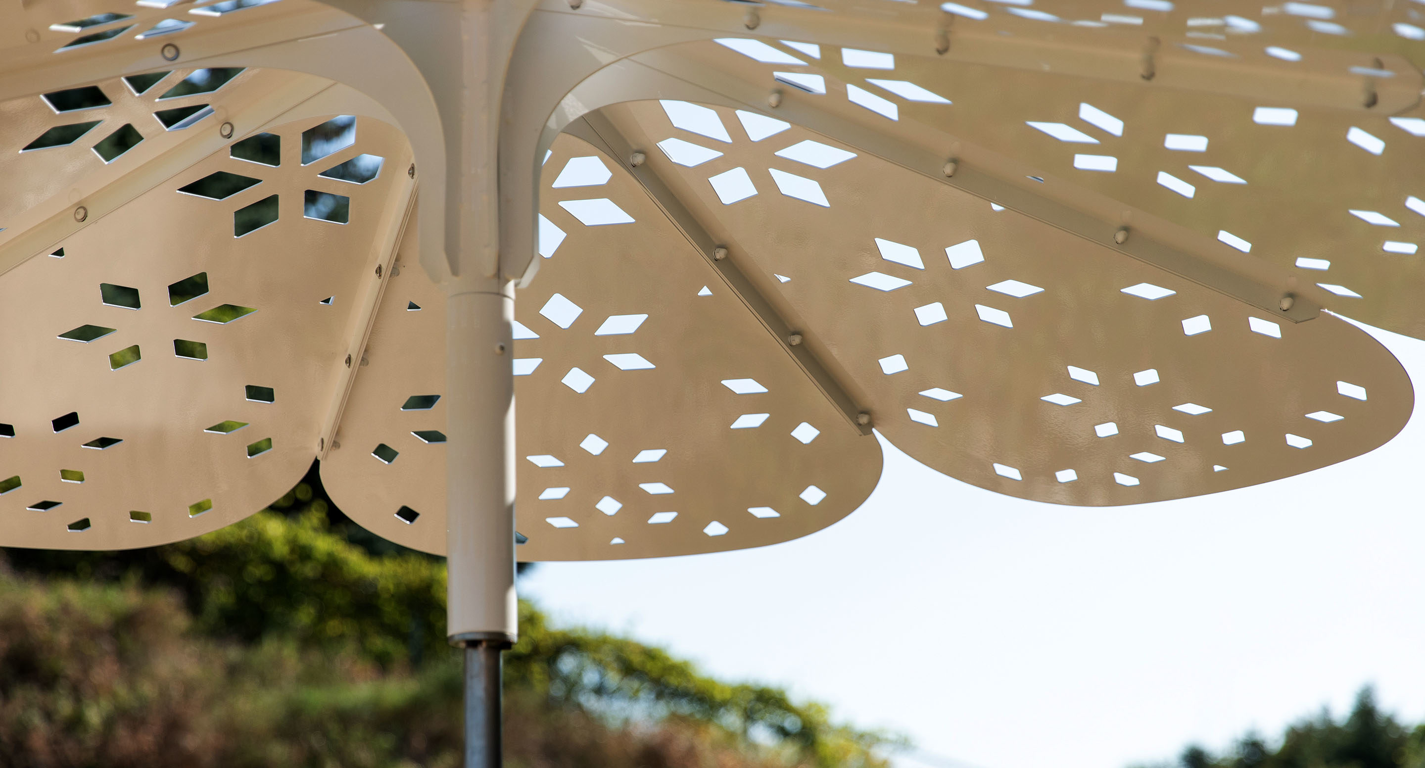 Close-up view of a white, metal, flower-shaped sunshade with geometric cut-out patterns, photographed outdoors against a blurred natural background.