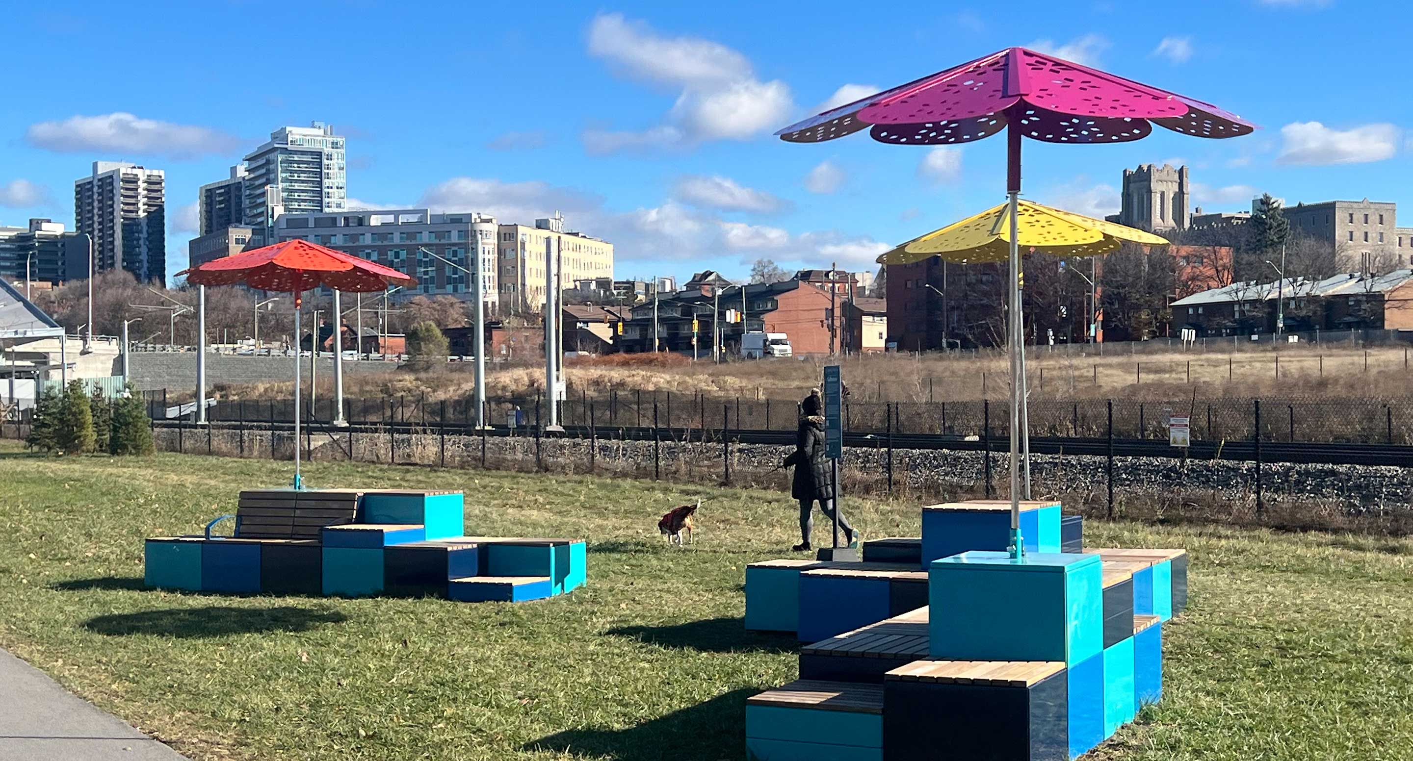 Colorful geometric benches with large metal umbrellas stand on grass near a path, with buildings, a statue, a dog, and blue sky in the background.