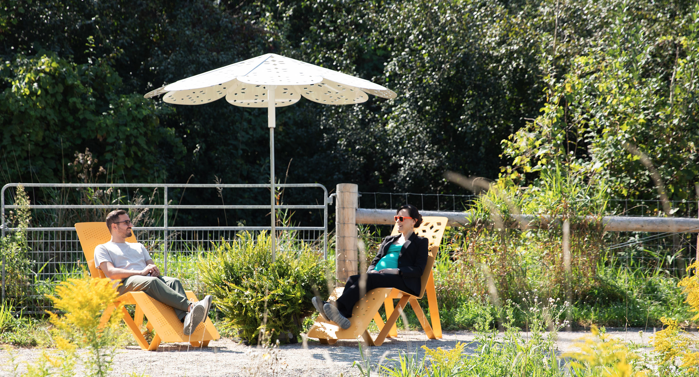 maglin_mum-3200-roma-umbrella-2 Deux personnes sont assises sur des chaises longues en bois sous un parasol blanc dans un espace extérieur avec de la verdure et une clôture métallique en arrière-plan.