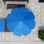 Vue aérienne d'un grand parasol bleu ombrageant un coin salon sur un patio en pierre, avec à proximité des bancs, des tables, des plantes et une place de parking pour handicapés partiellement visible.