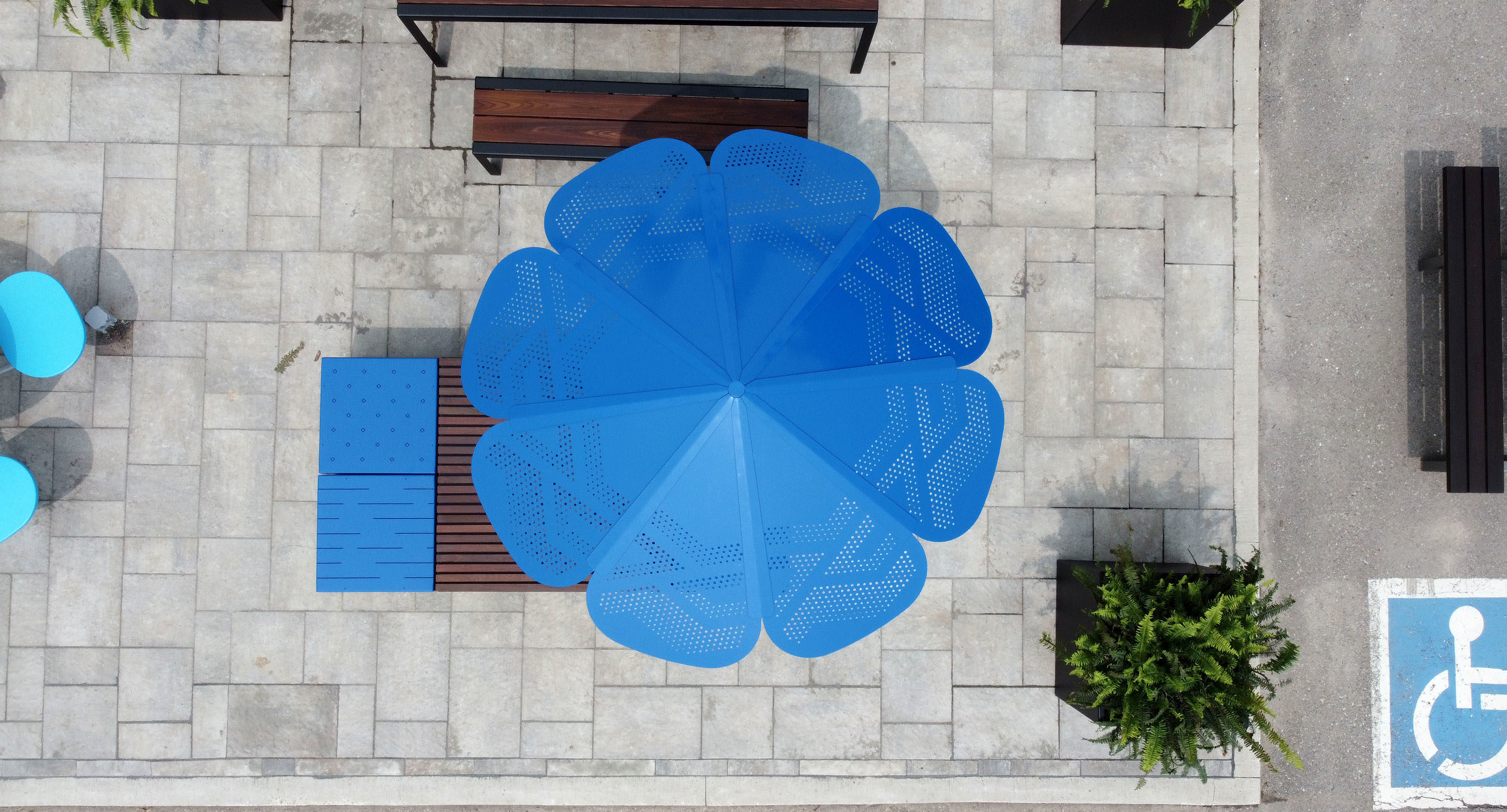 maglin_mum-3200-roma-umbrella-3 Vue aérienne d'un grand parasol bleu ombrageant un coin salon sur un patio en pierre, avec à proximité des bancs, des tables, des plantes et une place de parking pour handicapés partiellement visible.