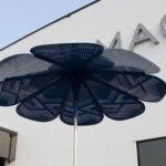 A large blue, flower-shaped metal structure with perforated patterns stands outside a modern building with partial signage visible.