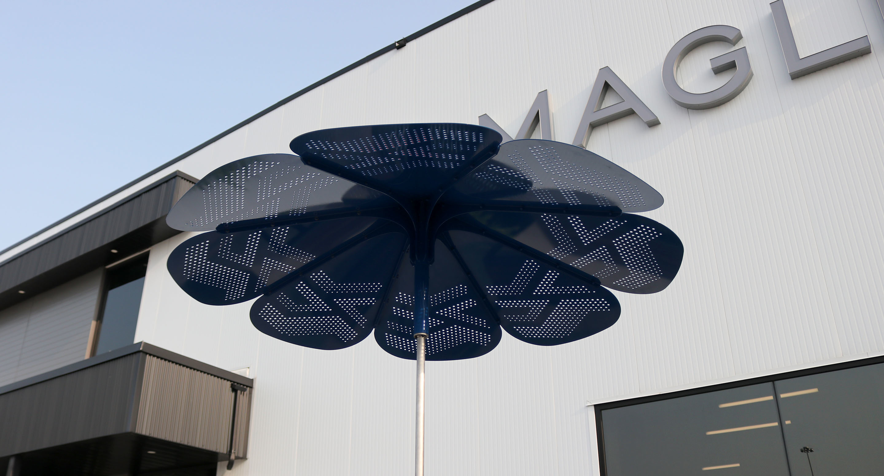 maglin_mum-3200-roma-umbrella-4 Une structure métallique bleue en forme de fleur se dresse devant un bâtiment moderne blanc dont la façade laisse entrevoir de grandes lettres.