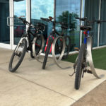 Four bicycles are parked in a metal bike rack outside a building with glass doors and windows, next to a patch of artificial grass.