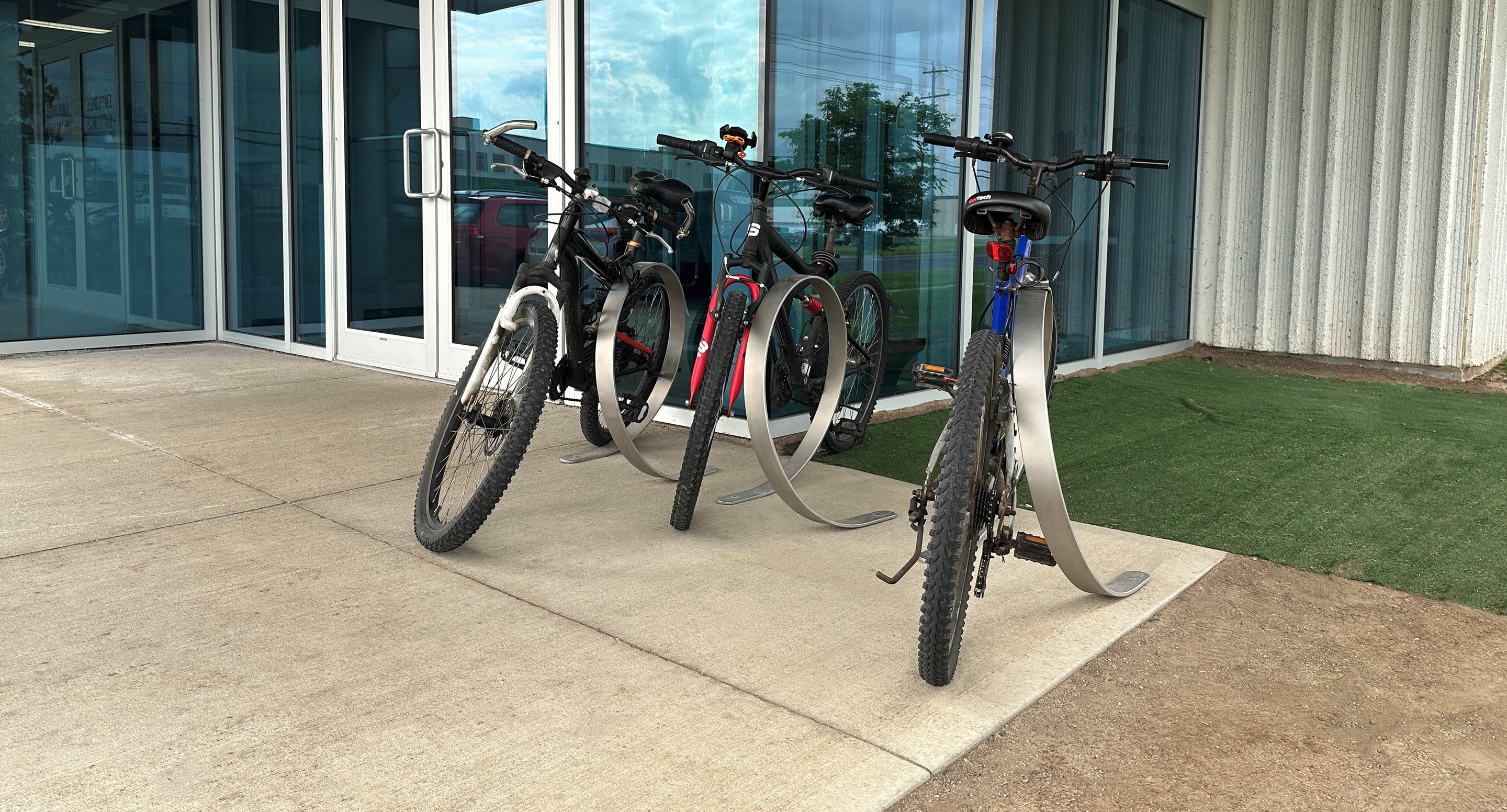 Four bicycles are parked in a metal bike rack outside a building with glass doors and windows, next to a patch of artificial grass.