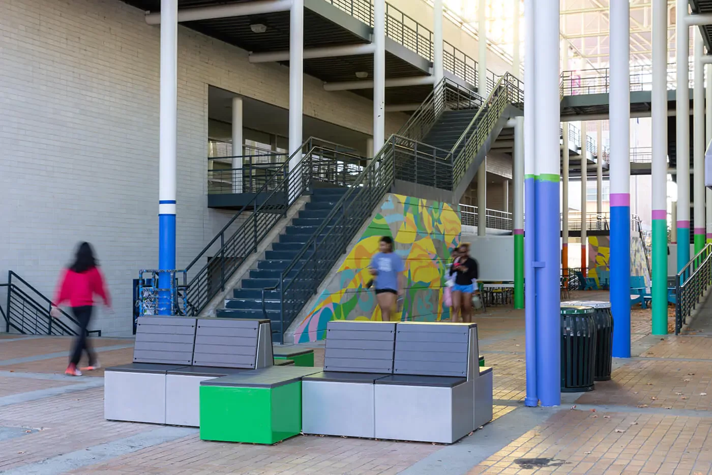 Espace extérieur avec des bancs modernes, des colonnes peintes en couleur et un escalier ; quelques personnes se promènent dans l'espace ouvert.