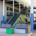 Open indoor public space with modern benches, mural on stair wall, several people walking, and colorful pillars.