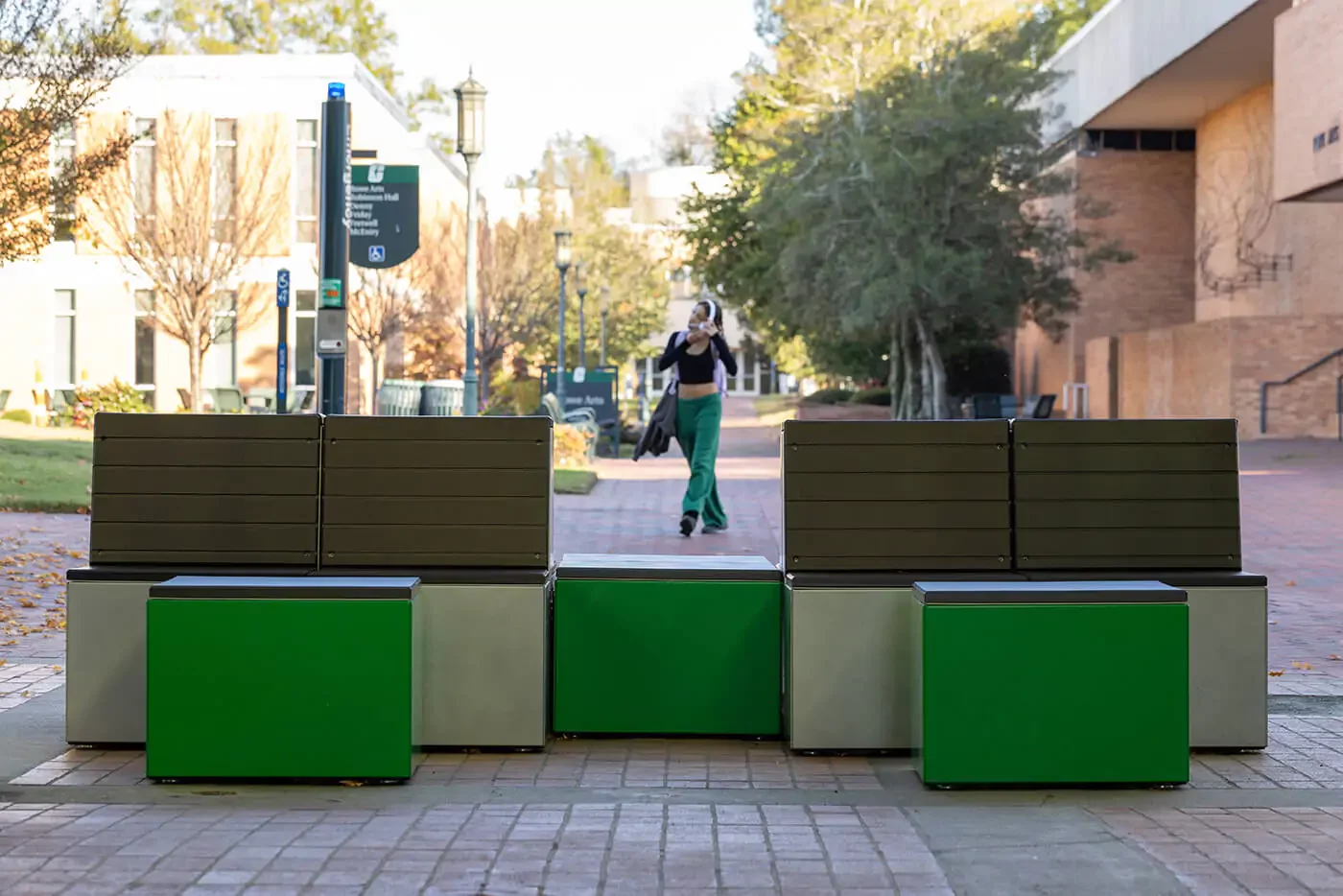A woman walks on a brick walkway past modern green and brown benches on a college campus, with trees and buildings in the background.