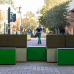 A woman walks on a brick walkway past modern green and brown benches on a college campus, with trees and buildings in the background.