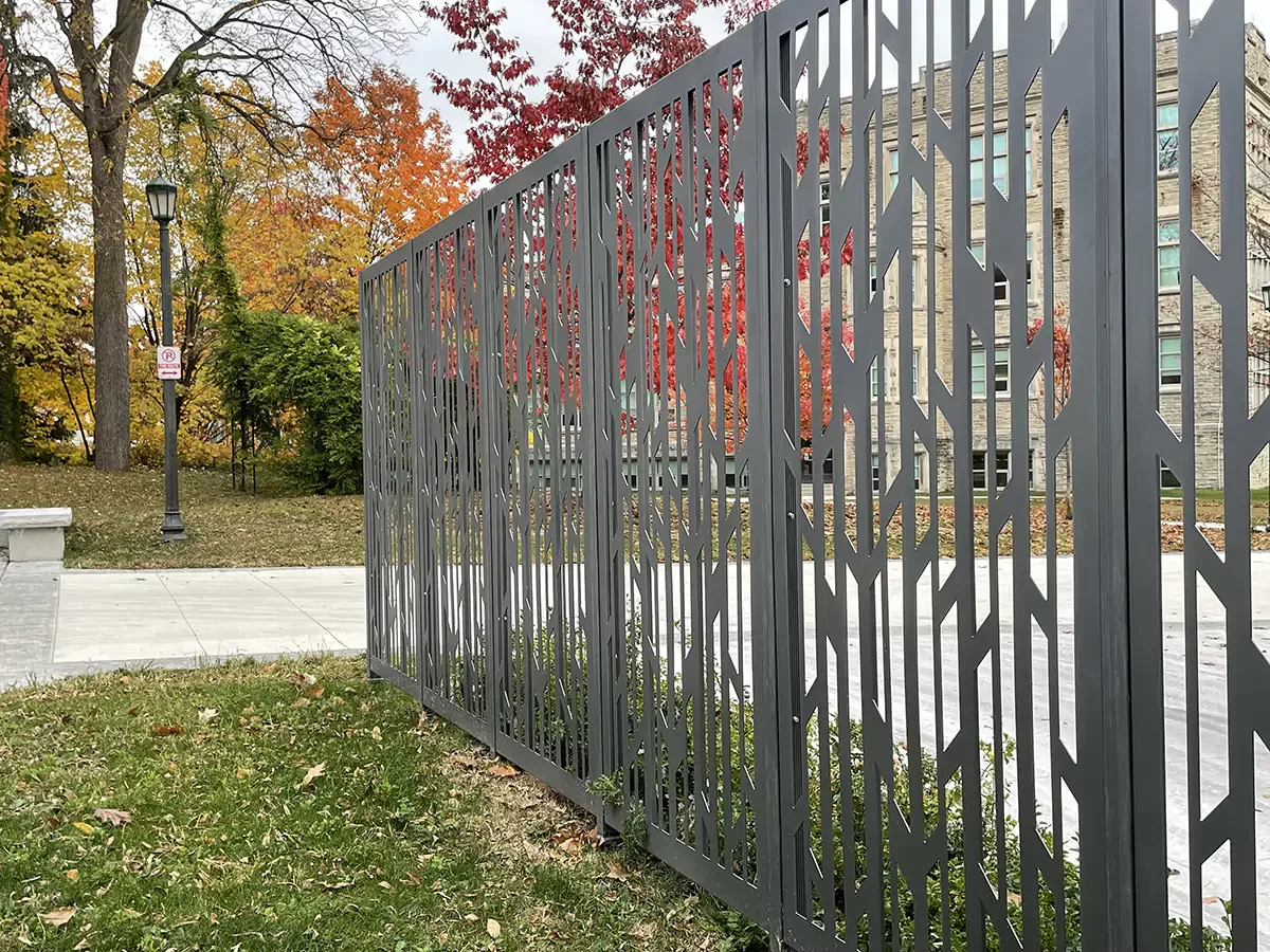 A decorative metal fence with a geometric pattern stands beside a sidewalk and grassy area; autumn trees and a building are in the background.