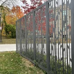 A decorative metal fence with a geometric pattern stands beside a sidewalk and grassy area; autumn trees and a building are in the background.