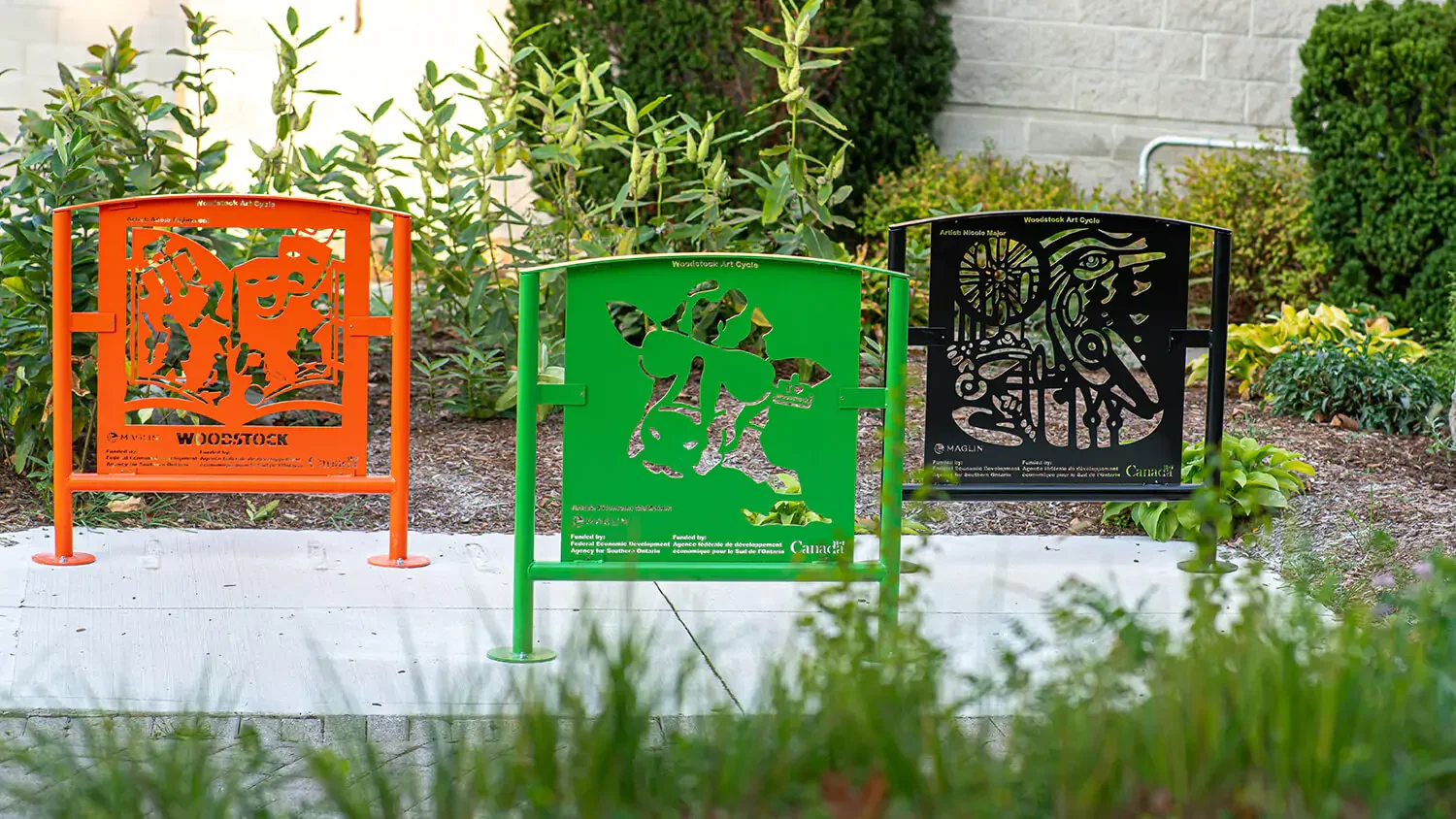Three colorful metal benches—orange, green, and black—each with intricate cut-out designs, are placed on a sidewalk in a landscaped garden area.