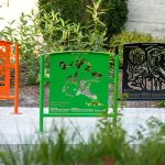 Three colorful metal benches—orange, green, and black—each with intricate cut-out designs, are placed on a sidewalk in a landscaped garden area.