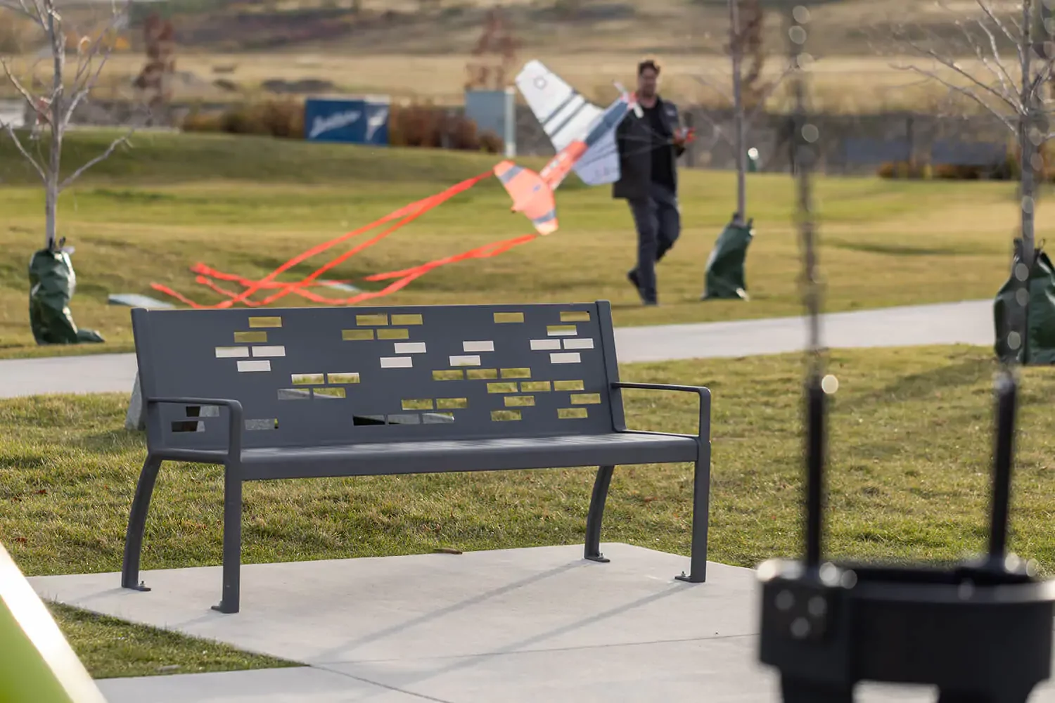 A gray metal park bench sits on a sidewalk with grass and young trees nearby; a person in the background carries a large model airplane.